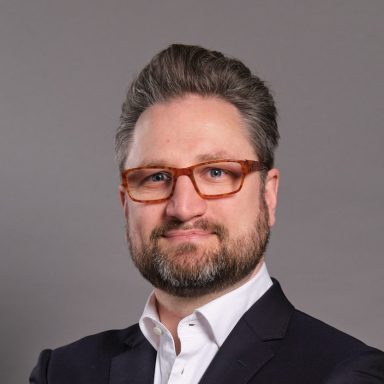 A man with glasses and a beard, wearing a suit, smiling confidently.