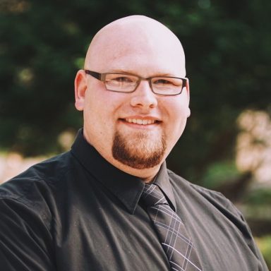 Smiling man with a bald head and glasses, wearing a black shirt and tie, outdoors.